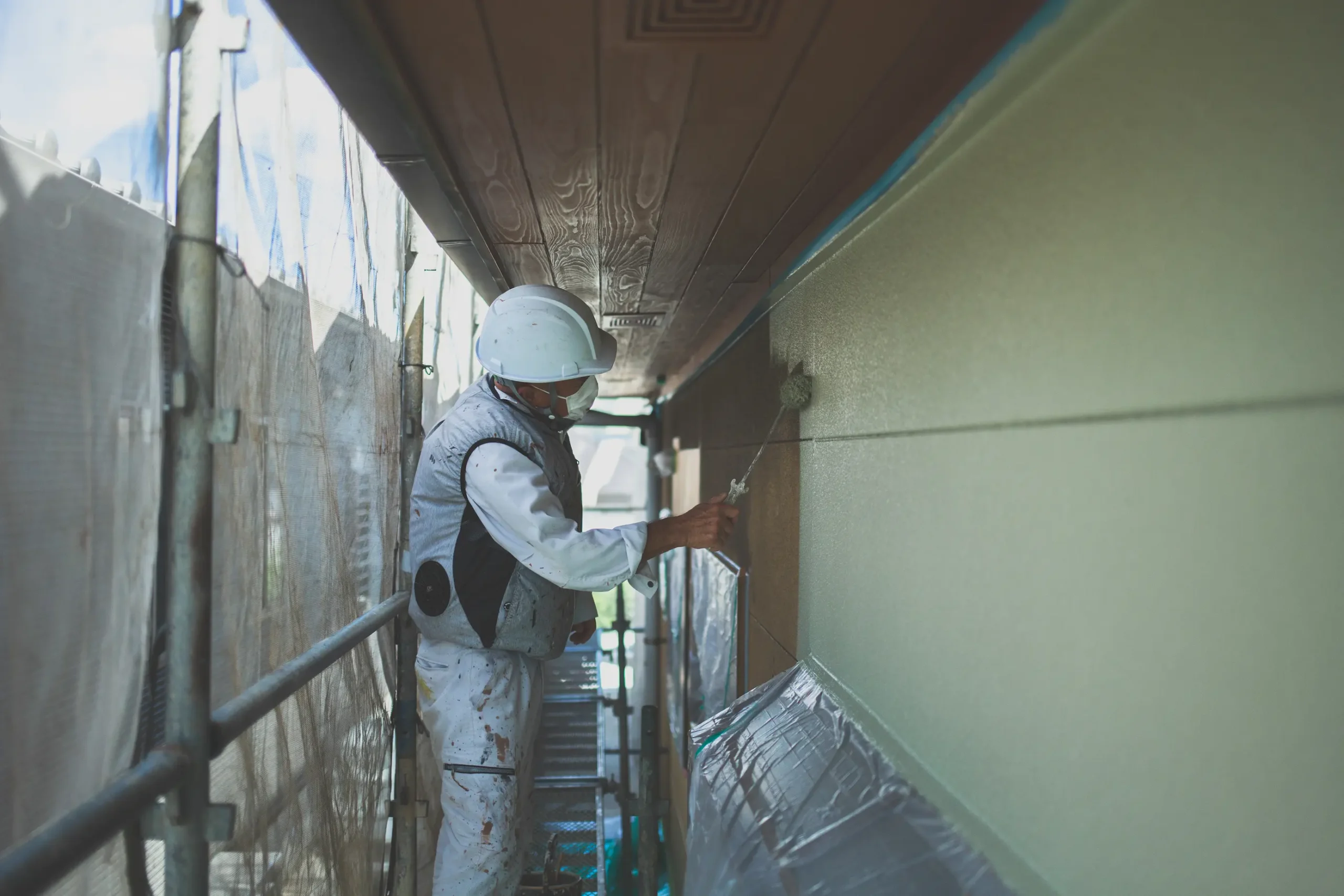 建物の外壁をローラーで丁寧に塗装する職人。安全対策としてヘルメットとマスクを着用し、足場の上で慎重に作業を進めている。品質の高い施工が多くのお客様から評価されている。
